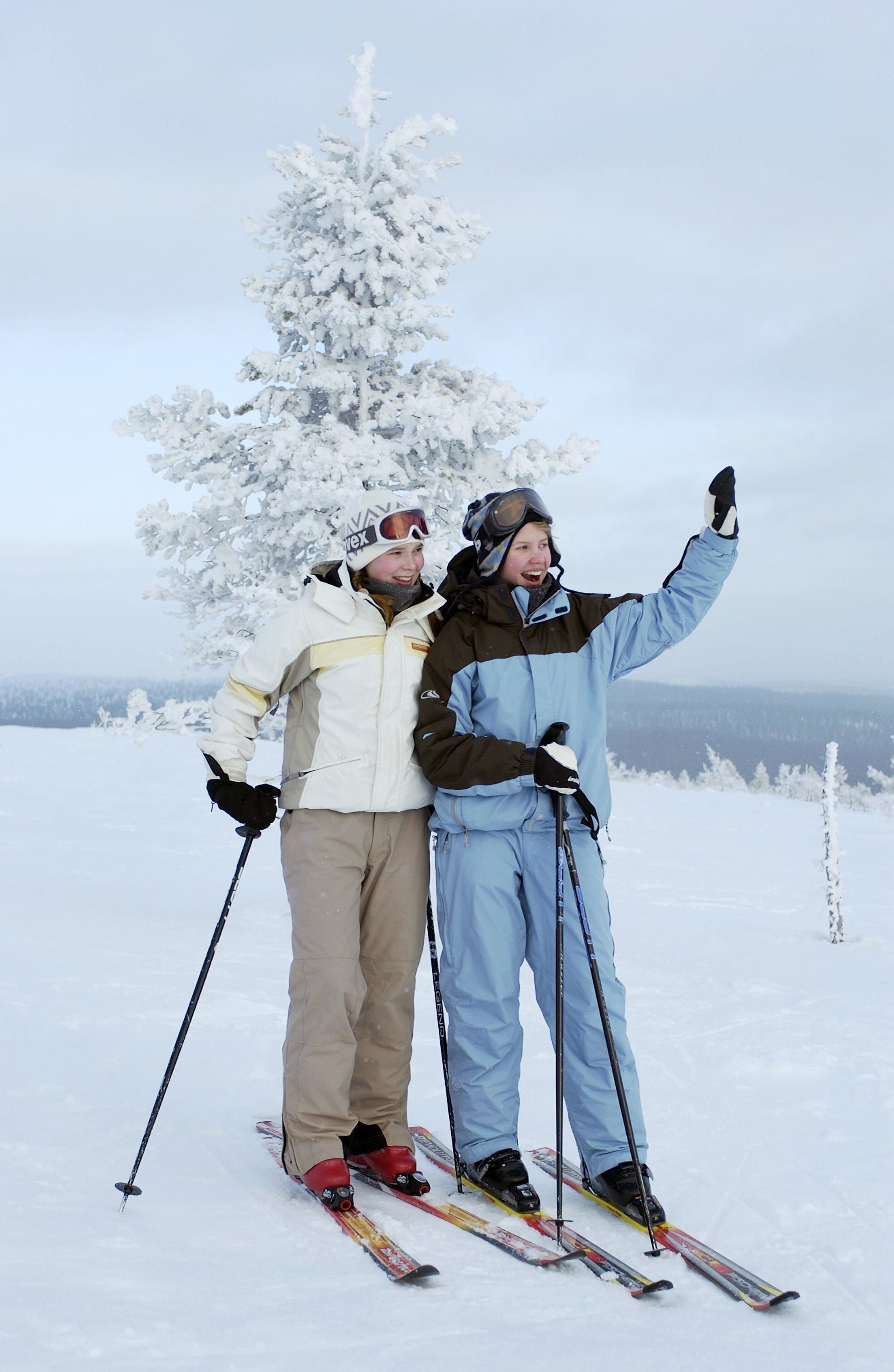 Two people skiing on a snowy slope with a snow-covered tree in the background.