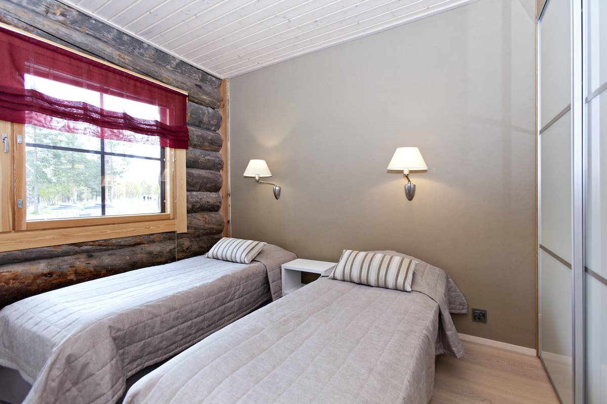 A room with two beds, a window with a red curtain, and two wall-mounted lamps.