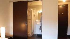Una habitación de hotel con cama, baño con ducha y puerta que da a otra habitación.