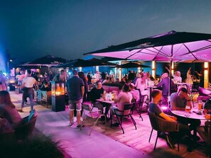 An outdoor dining area with tables and chairs, umbrellas, and a crowd of people.