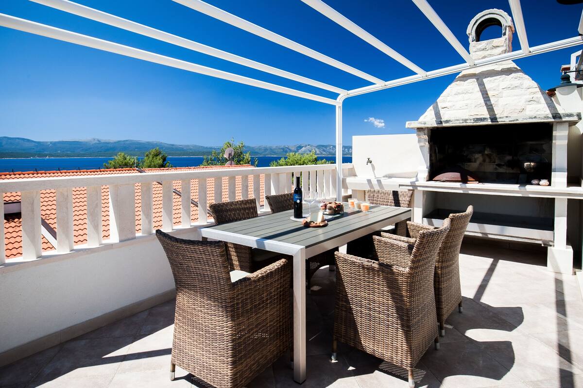 Eine Terrasse mit Tisch, Stühlen, einem Steinofen und Blick aufs Meer.
