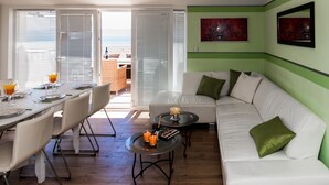 A modern living room with a white sofa, green cushions, a dining table set for four, and a view of the outdoors through large windows. - Mazuranic Apartments (Bol)