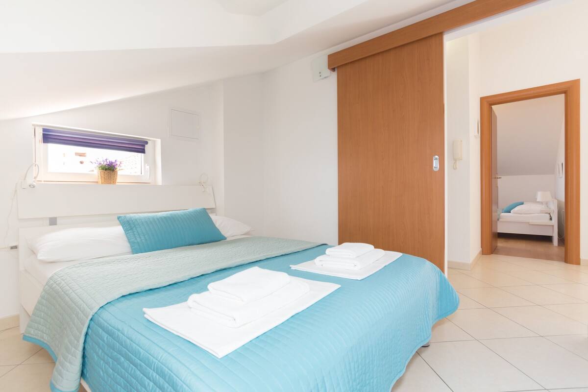 Ein ordentlich bezogenes Bett mit blauer Bettdecke und weißen Kissen, eine Holztür und ein kleines Fenster mit einem Blumenkorb.