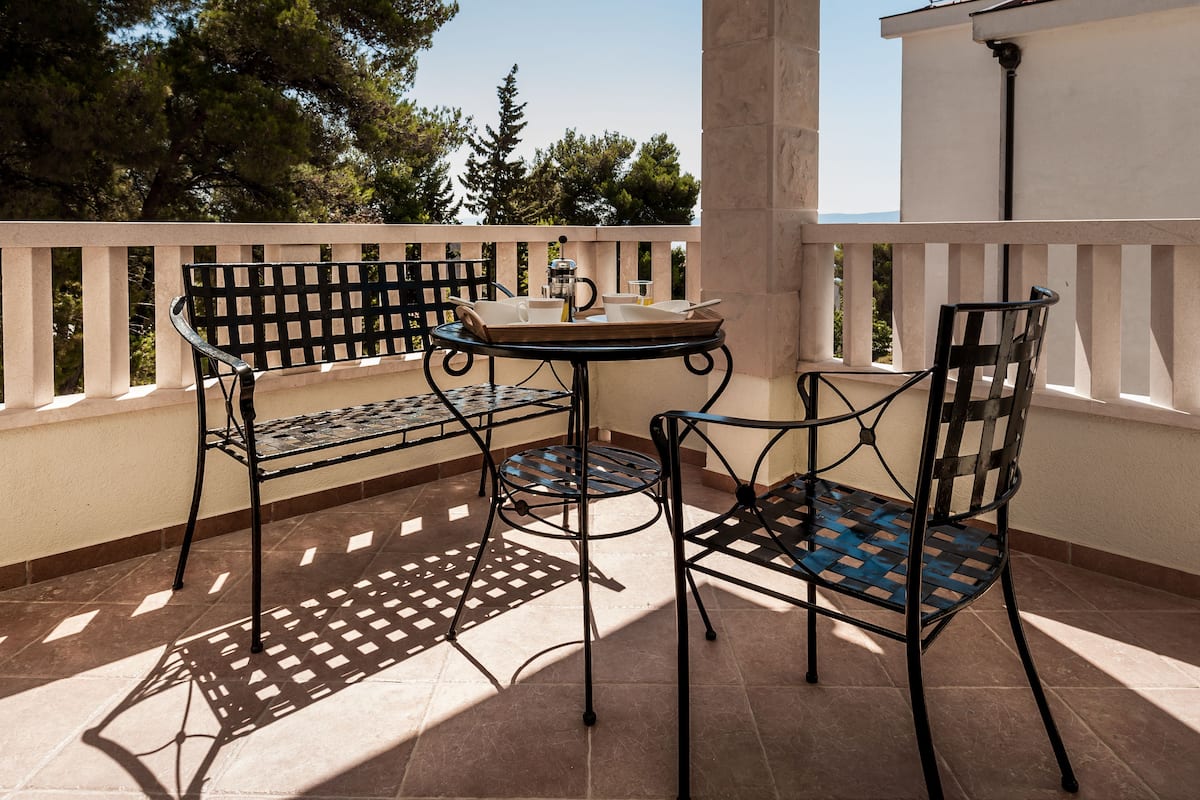 Eine Terrasse im Freien mit einem runden Tisch und zwei Stühlen, ein kleiner Tisch mit einer Tasse und einer Flasche, sowie Blick auf Bäume und ein Gebäude im Hintergrund.