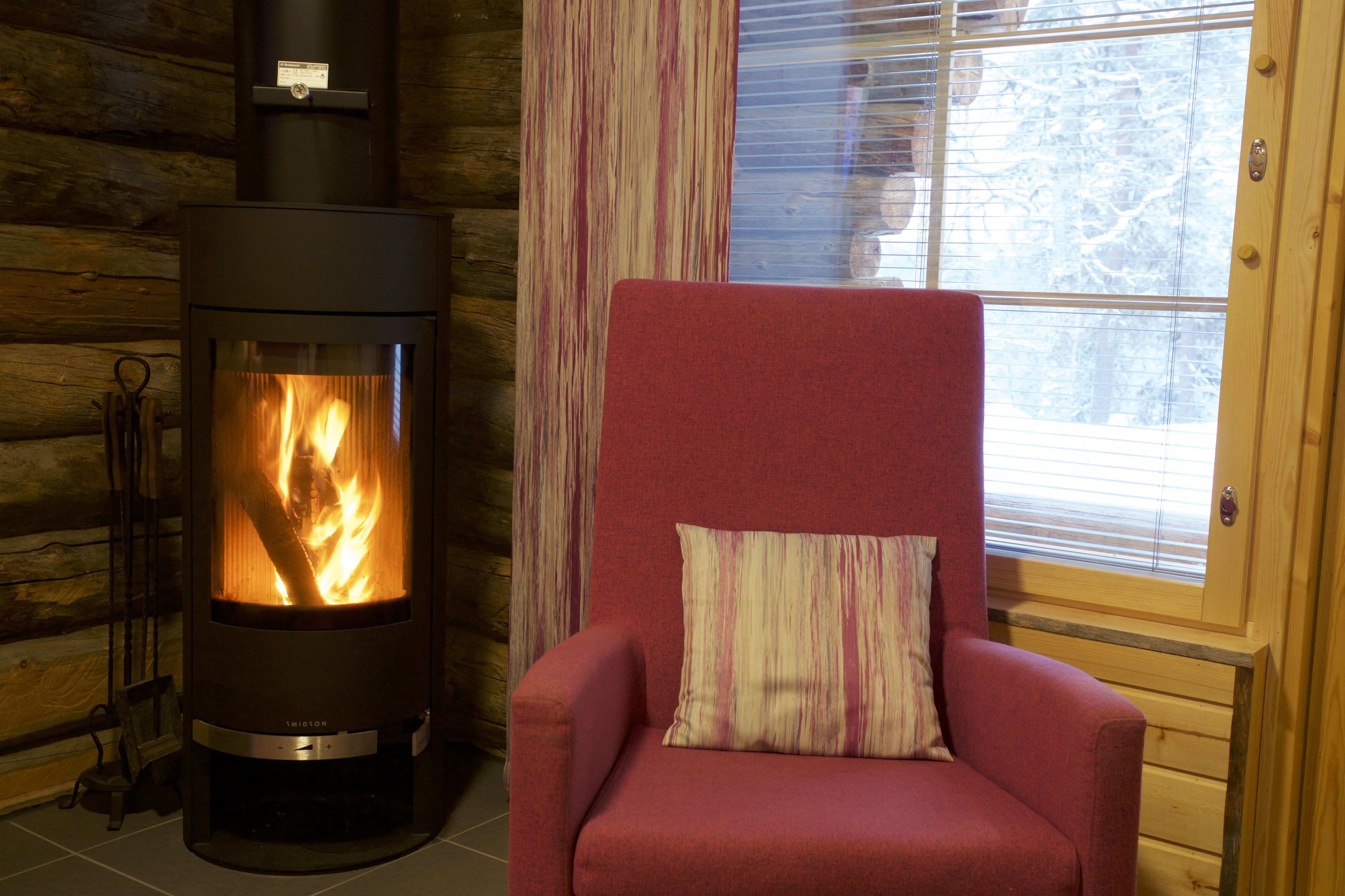 A log cabin interior with a wood-burning stove, a red armchair with a striped pillow, and a window with blinds.