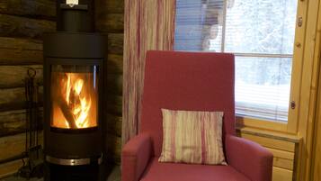 A log cabin interior with a wood-burning stove, a red armchair with a striped pillow, and a window with blinds.