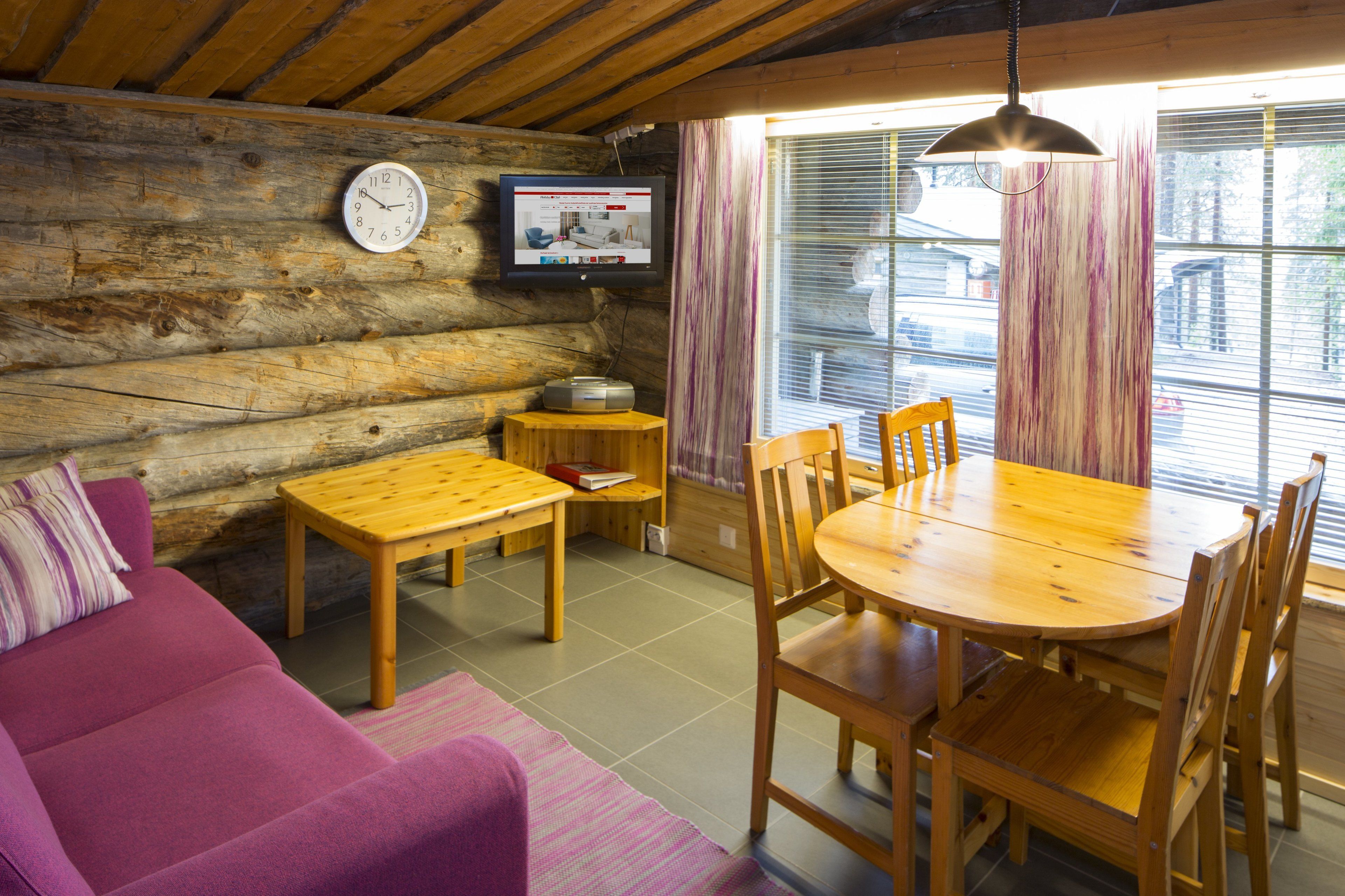 A cozy log cabin interior with a wooden dining table, chairs, a sofa, a television mounted on the wall, and a window with curtains.