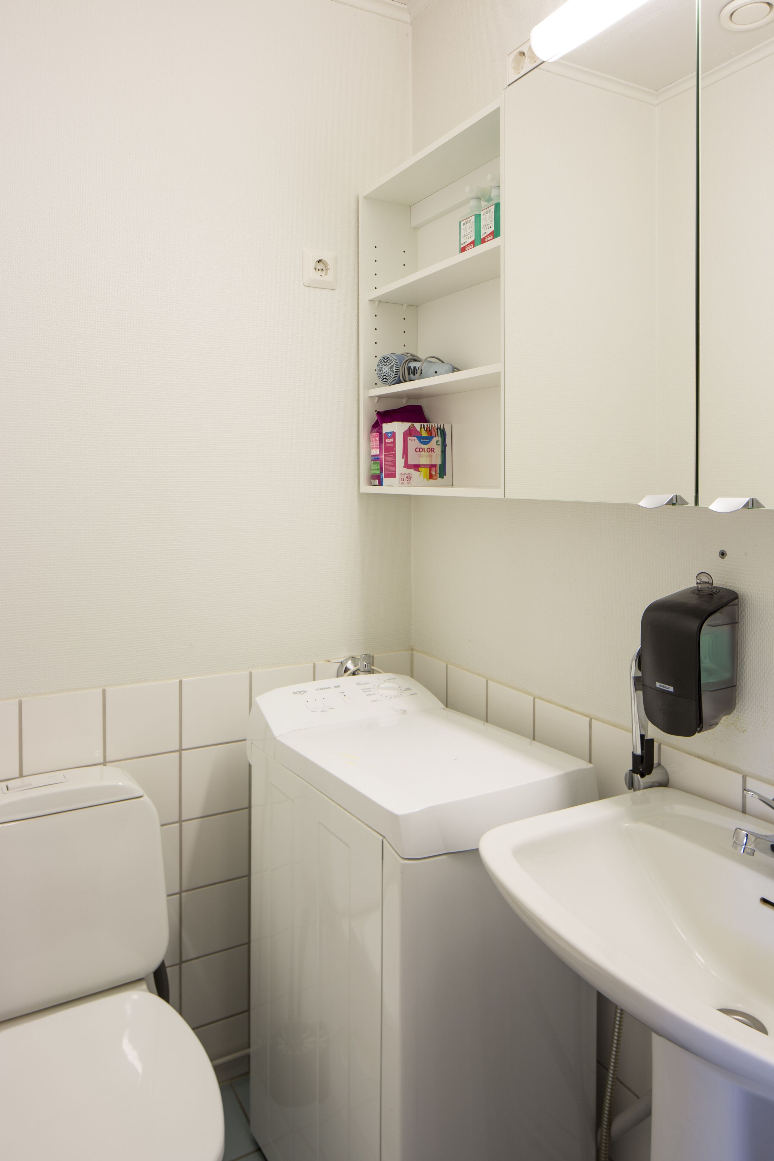 A bathroom with a toilet, a sink, and a mirror. There is a shelf with toiletries and a soap dispenser.