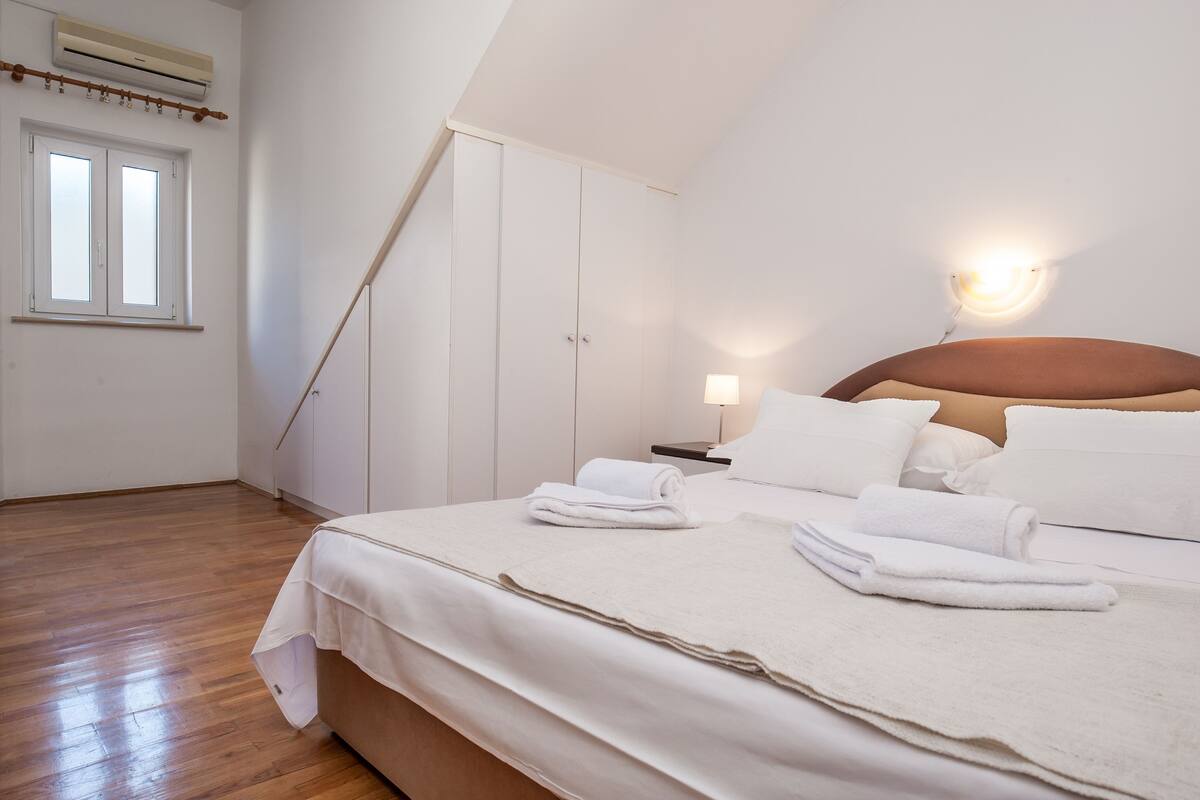 A bedroom with a bed, white linens, a nightstand, a lamp, and a window.