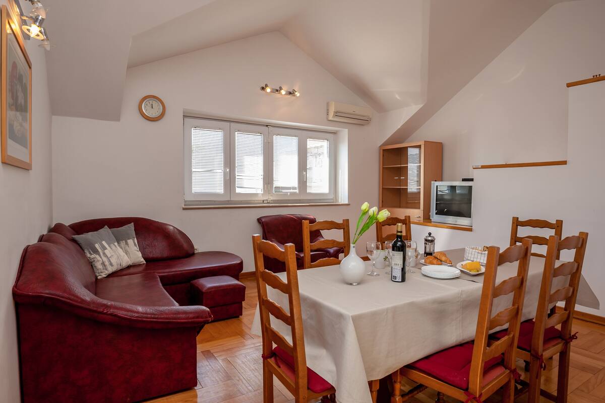 A dining area with a table set for four, a red leather sofa, wooden chairs, and a clock on the wall.