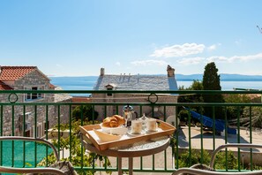 A balcony with a tray of croissants, coffee, and tea, overlooking a pool and a scenic view of the sea and mountains.