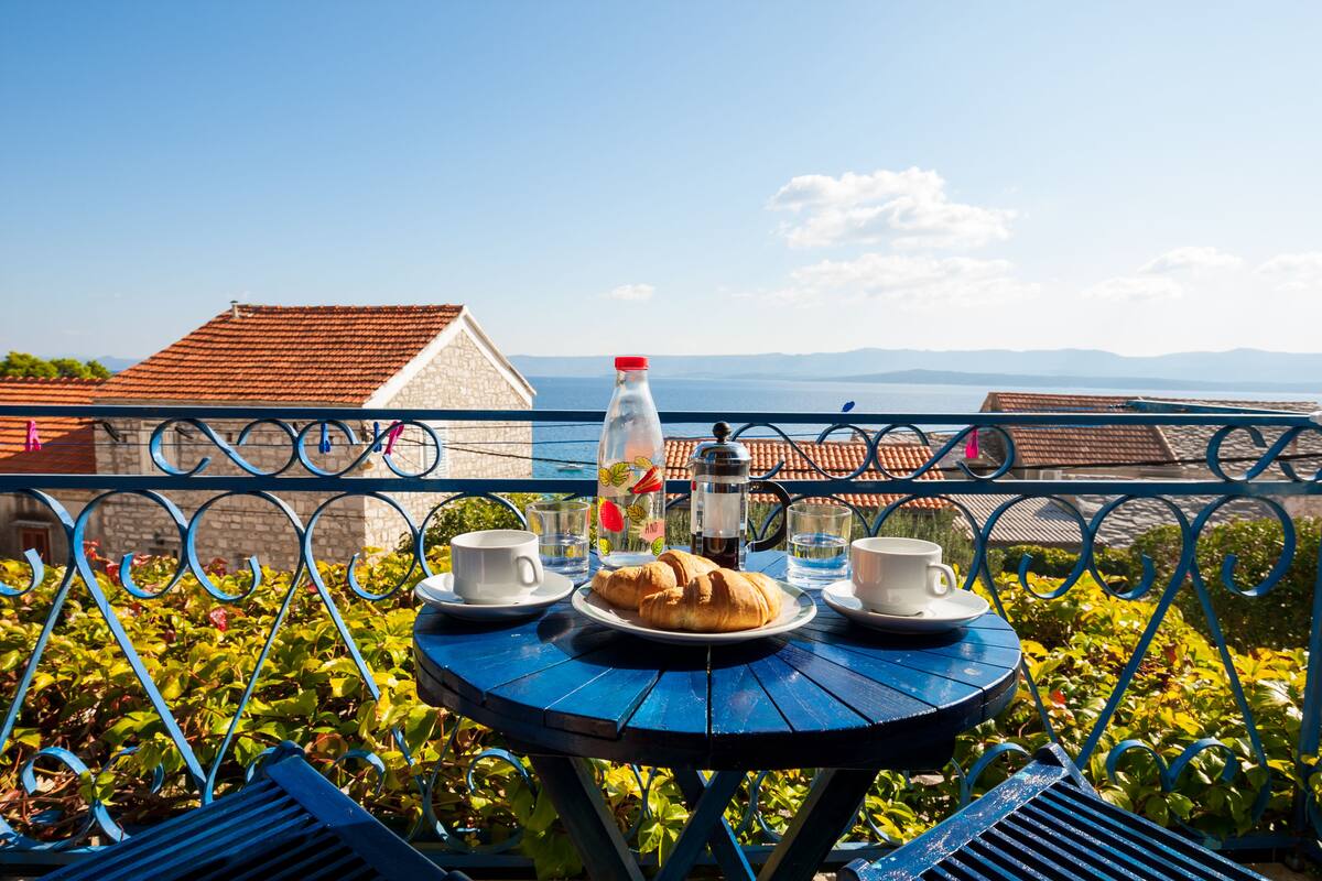 A round table with a croissant, two cups, and a bottle of water, set against a scenic backdrop with a view of the sea and distant mountains.
