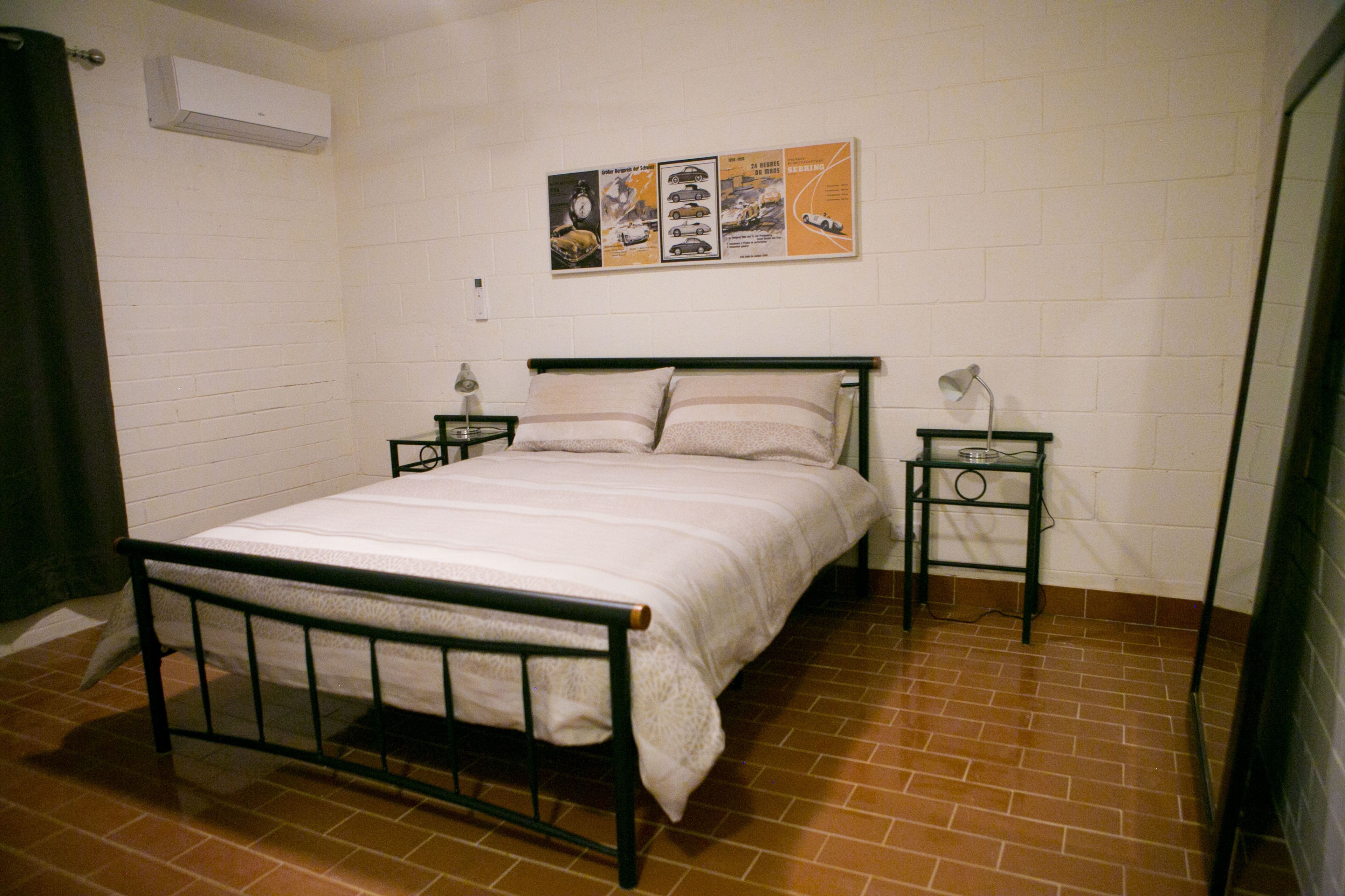 Una cama individual con estructura metálica, dos mesitas de noche y un aire acondicionado instalado en la pared.