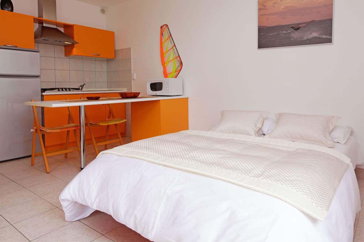 A bedroom with a bed, a desk, and a chair. There is a kitchen area with a stove and a microwave.