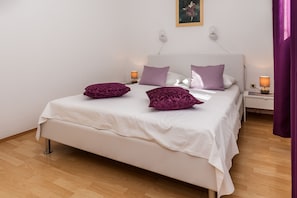 A neatly made bed with white bedding and purple pillows, a bedside table with a lamp, and a wall-mounted light fixture. - Ratac Apartments (Bol)