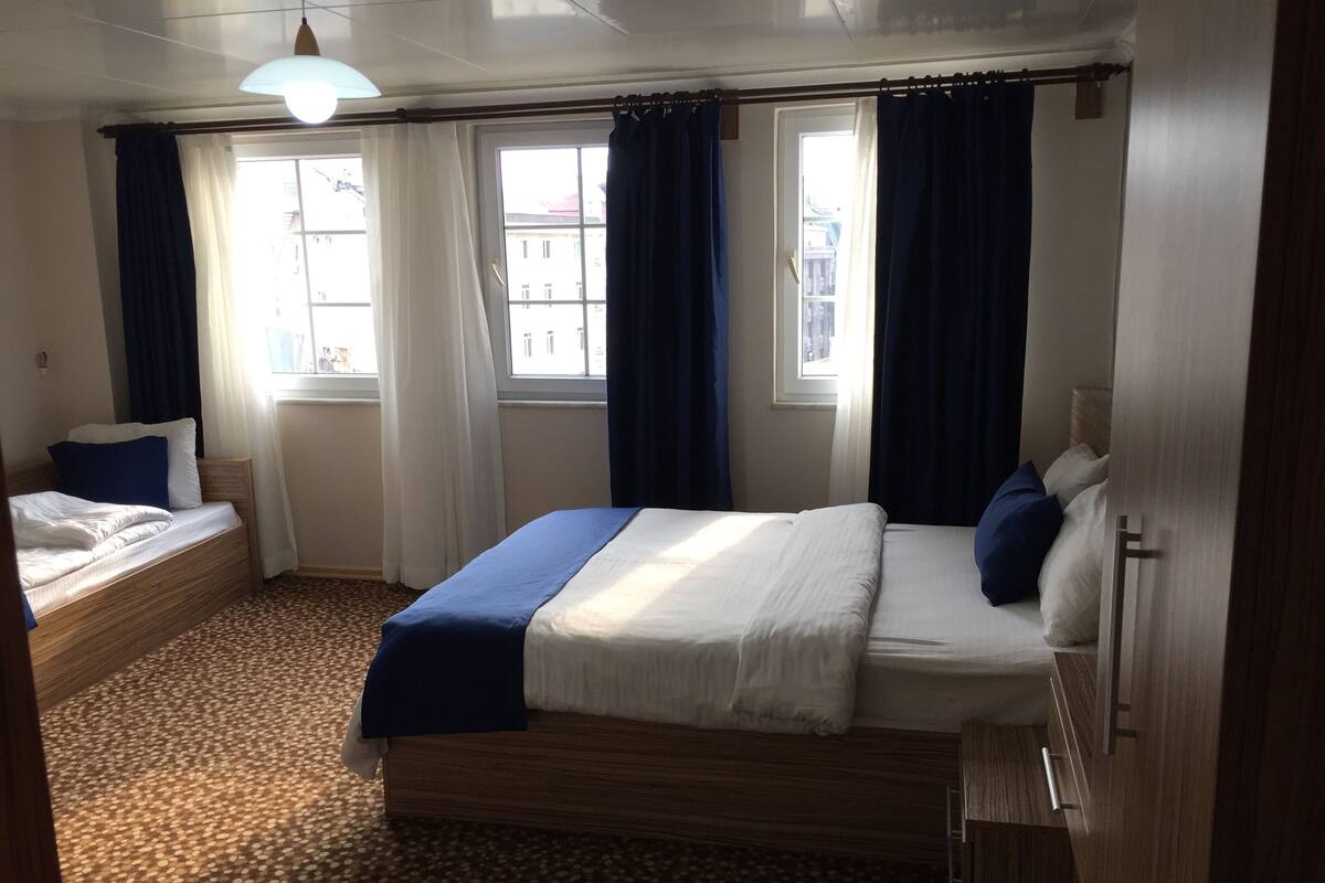 A hotel room with a large bed, a smaller bed, two windows with curtains, a patterned carpet, and a ceiling light.