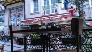 Ett utomhusområde med bord och stolar, utsikt över en byggnad med skylten ”ASLAN HOME APART”, och en butik med skylten ”ASLAN GIFT SHOP”.