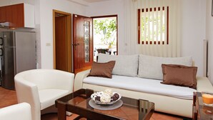Una sala de estar con un sofá, dos sillones blancos, una mesa de centro de vidrio con un cuenco decorativo, un refrigerador y una ventana con persianas.