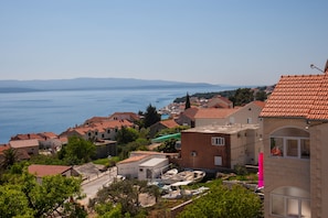 A coastal town with terracotta-roofed buildings, a clear view of the sea, and a prominent pink flag.