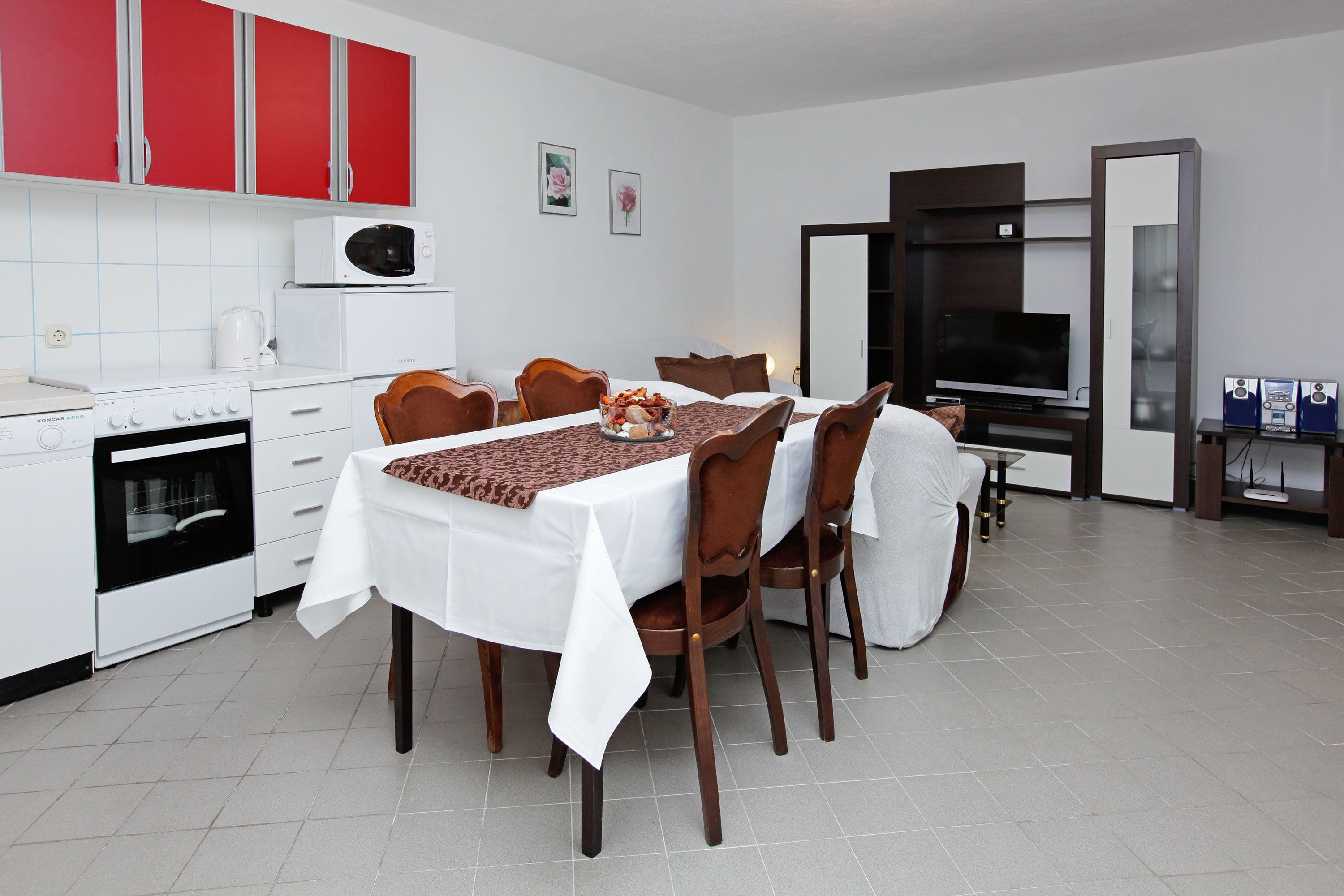 Une cuisine moderne avec des meubles blancs, un four à micro-ondes et une cuisinière. On y trouve une table à manger avec des chaises, et un téléviseur en arrière-plan.