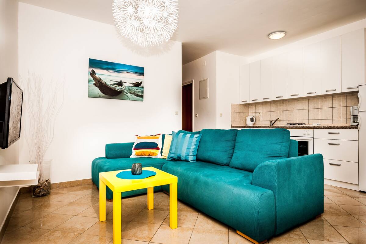 Ein modernes Wohnzimmer mit einem türkisfarbenen Sofa, einem gelben Couchtisch und einer Küchenzeile mit weißen Schränken und einer gefliesten Spritzschutzwand.
