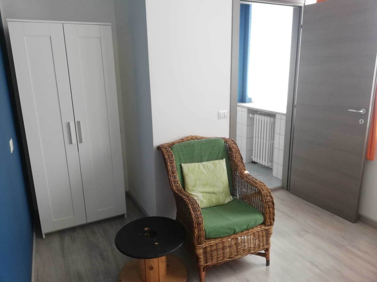 A room with a wicker chair, a small round table, a wardrobe, and a door leading to a balcony.