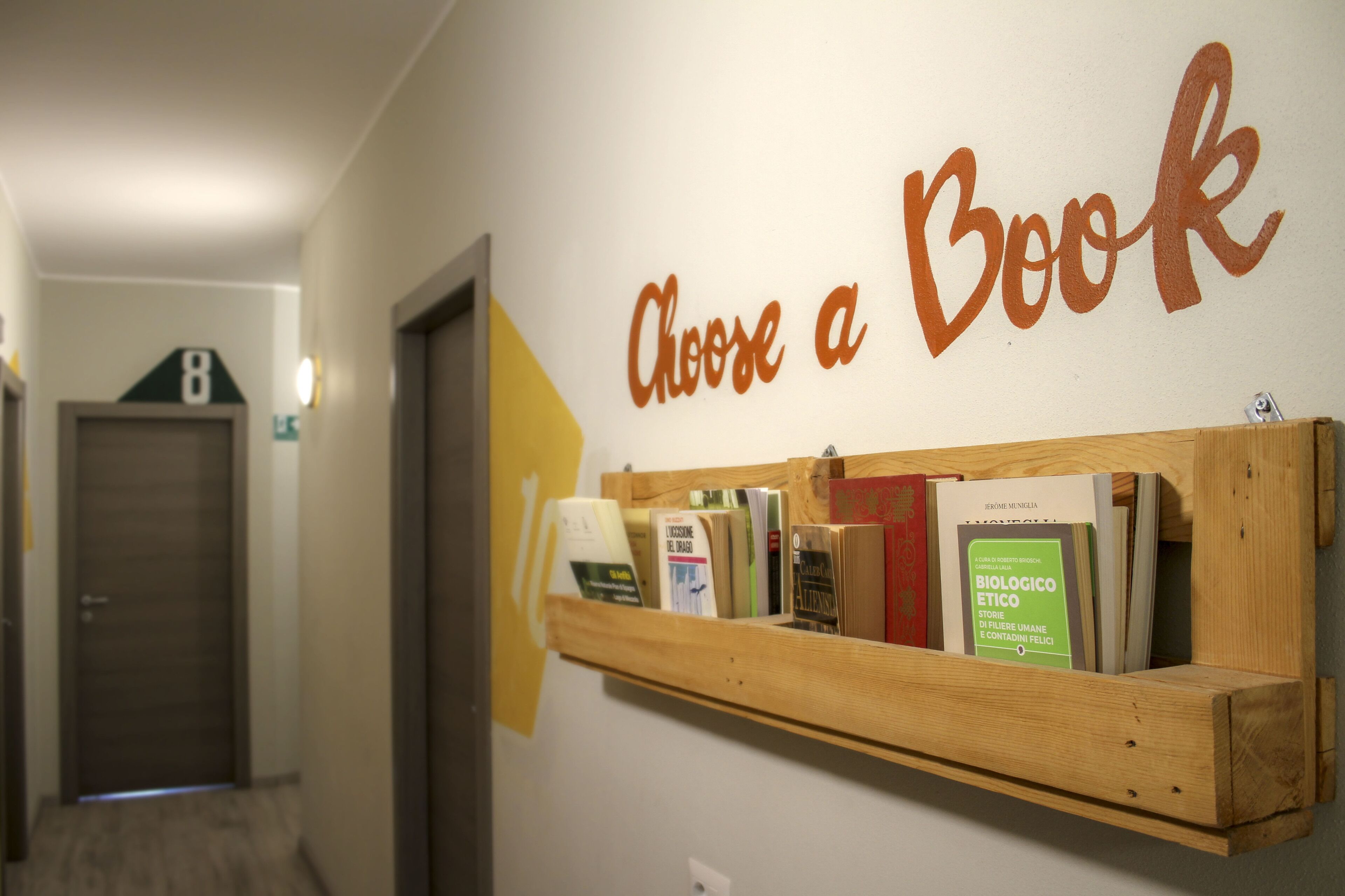 Un corridoio con una libreria in legno che espone libri e un cartello con la scritta 'Choose a Book'.