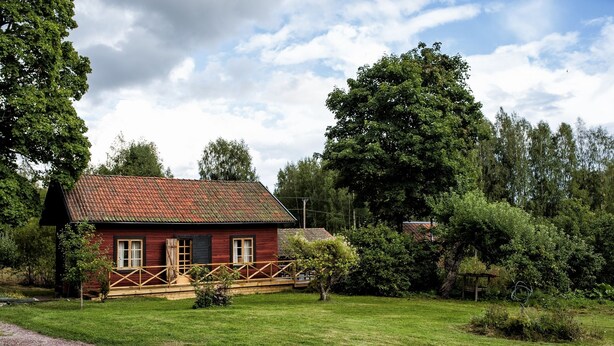 Une maison en bois rouge, avec un toit en tuiles, entourée d’une cour verdoyante et d’arbres.