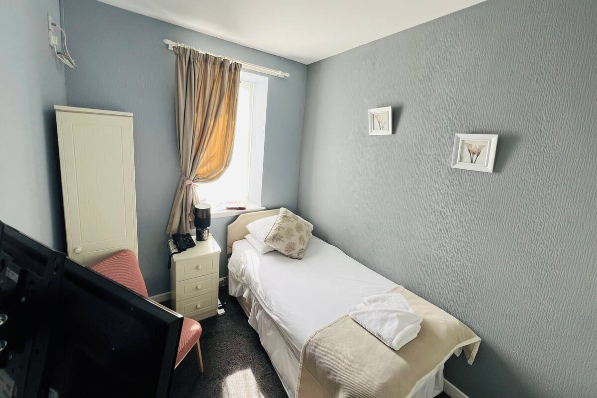 Una habitación de hotel con cama, mesita de noche, silla, armario y ventana con cortinas.