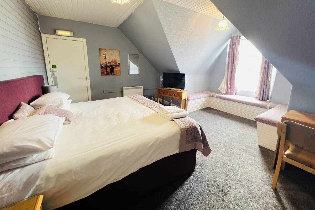Un dormitorio con cama, televisión, escritorio y ventana con cortinas.