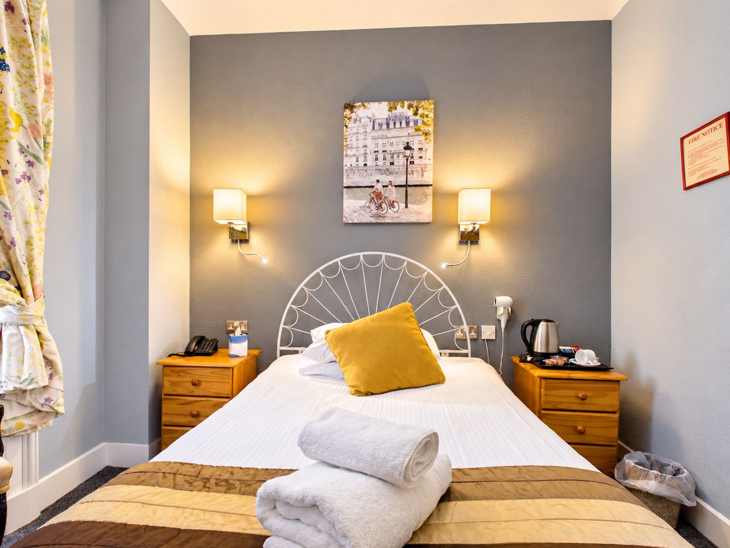 A hotel room with a bed, two bedside tables, a headboard, a painting on the wall, and a fire notice.