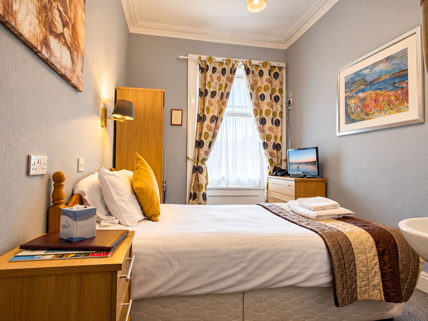 Una habitación de hotel con una cama, un escritorio con lámpara, un televisor y un cuadro en la pared.