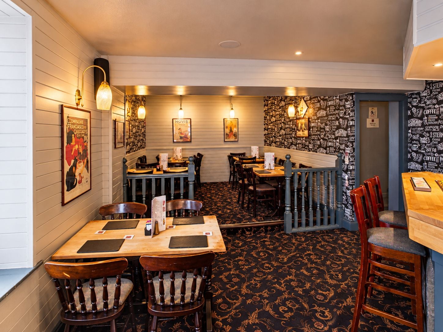 Ein Restaurantinneres mit Holztischen und -stühlen, Wandleuchten und einem gemusterten Teppich.