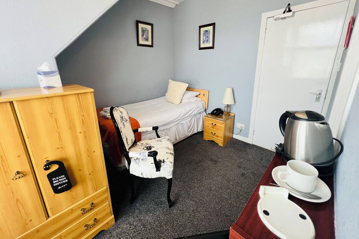 Una habitación pequeña y ordenada con una cama, un armario de madera, un escritorio con tetera y tazas, y una silla.