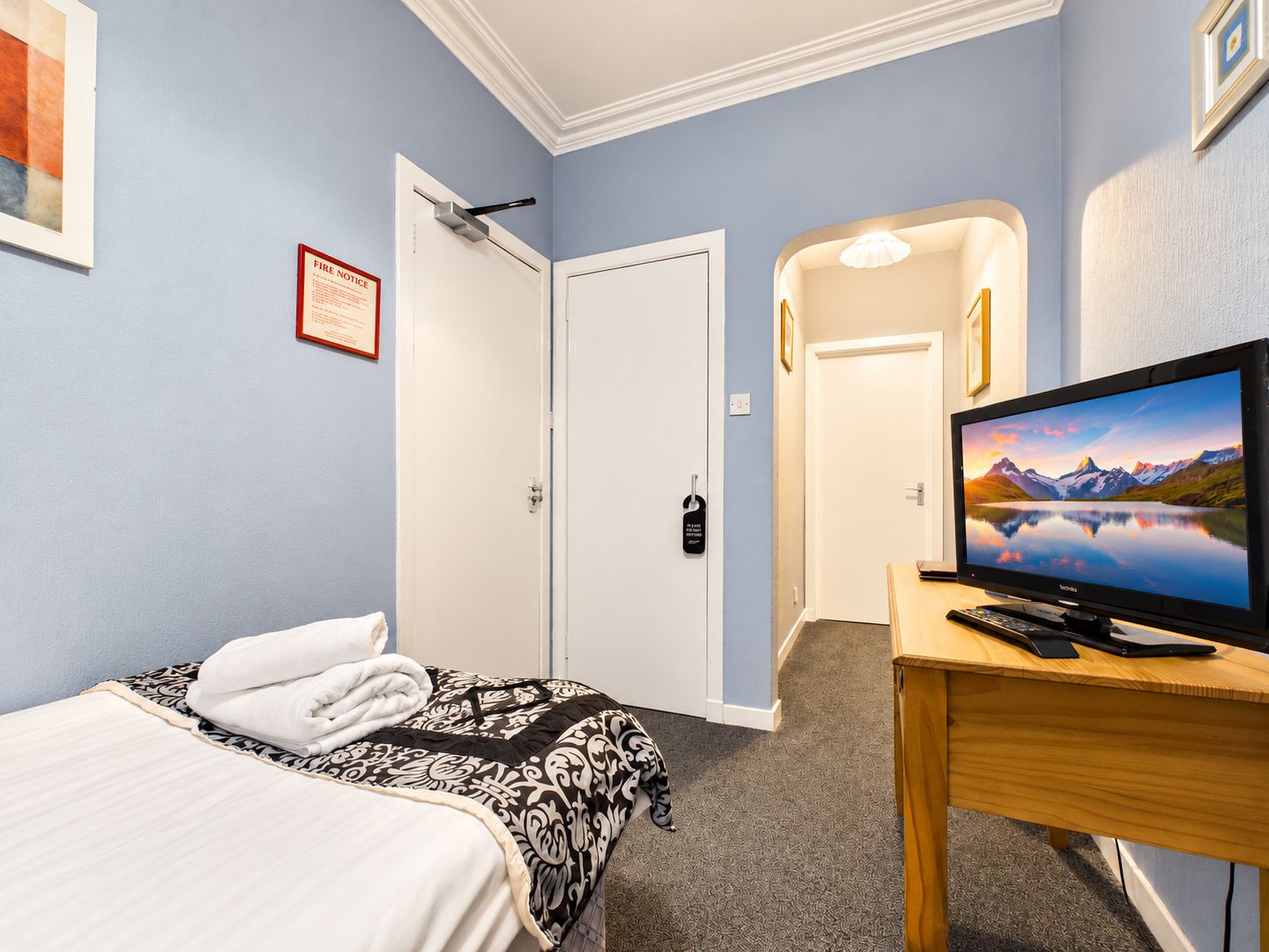 A hotel room with a bed, a television on a wooden table, a fire notice on the wall, and a door leading to another room.