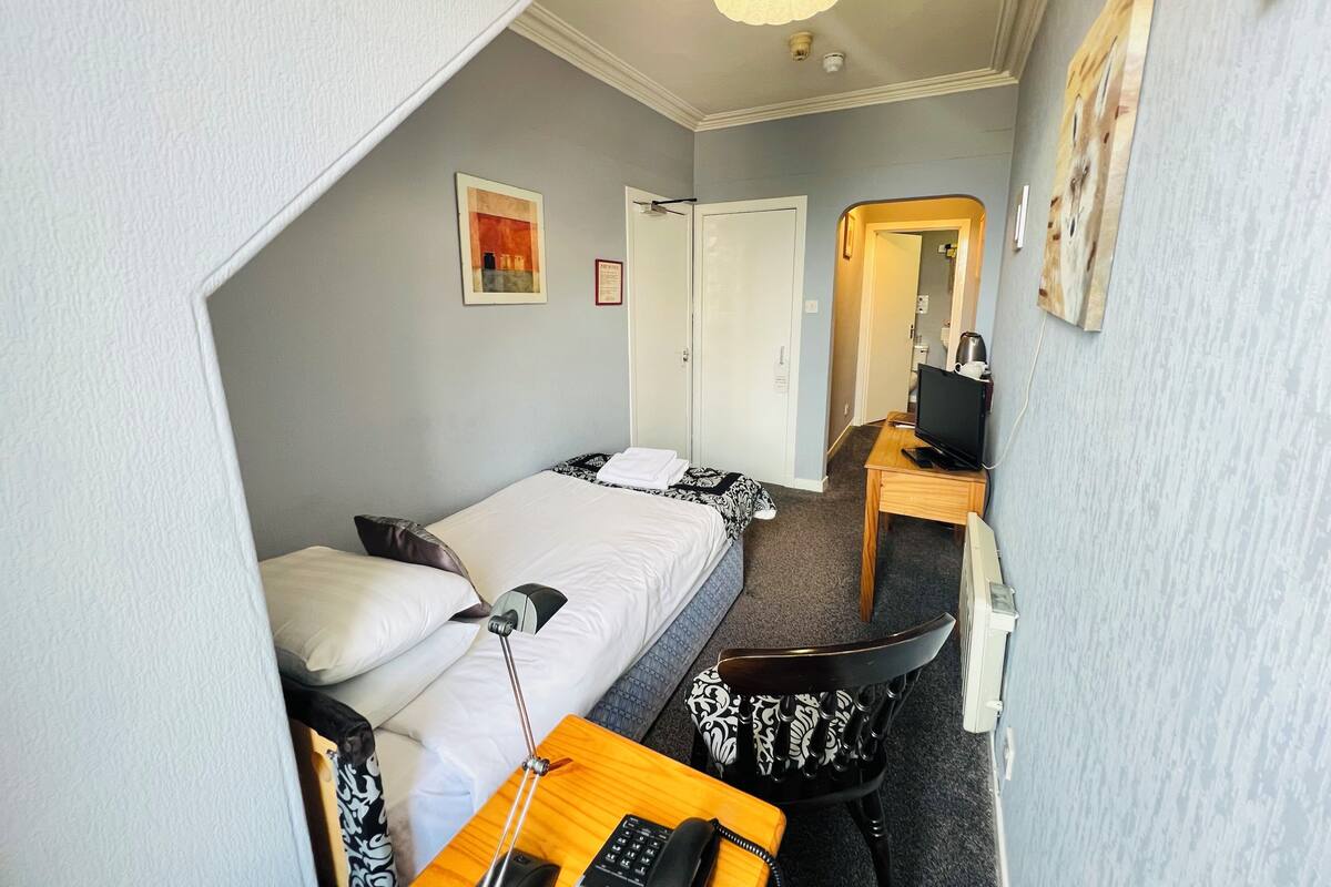Habitación de hotel compacta con cama, escritorio con computadora, una silla y una mesita de noche con lámpara.
