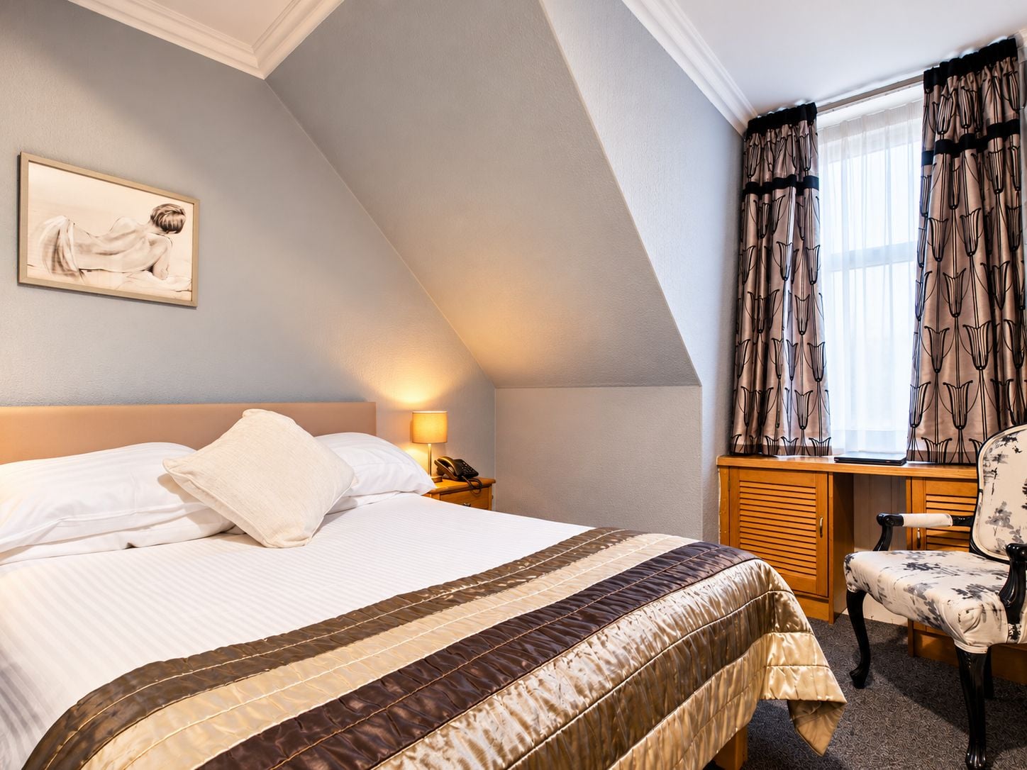 Una habitación de hotel con una cama, una mesita de noche con lámpara, una silla y una ventana con cortinas.