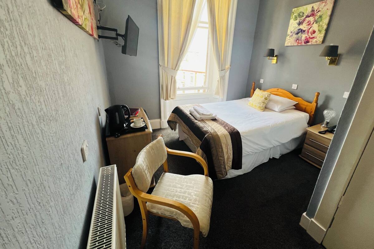 Una habitación de hotel con una cama, una silla, un escritorio con cafetera, una lámpara y un cuadro en la pared.