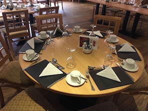 Una mesa redonda para comer dispuesta con tazas, platillos y cubiertos blancos, rodeada de sillas de madera.