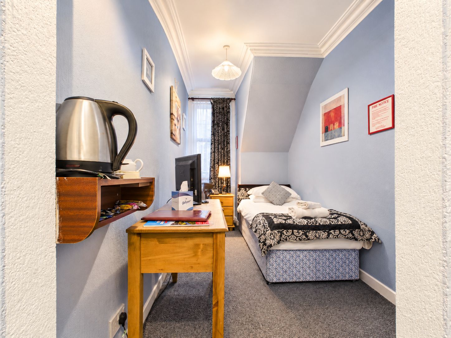 Een kleine hotelkamer met een bed, een bureau, een stoel, een nachtkastje en een wandplank.