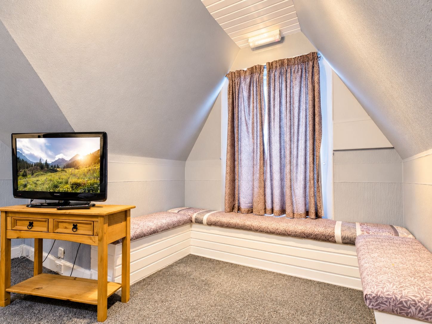 A cozy attic bedroom with a television on a wooden stand, a bench with cushions, and a window with patterned curtains.