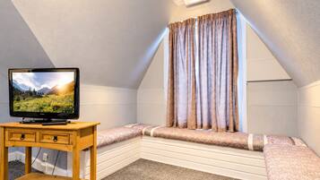 A cozy attic bedroom with a television on a wooden stand, a bench with cushions, and a window with patterned curtains.