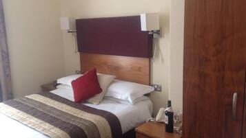 A hotel room with a bed, a bedside table with a bottle and glasses, a wooden headboard, and a wardrobe.