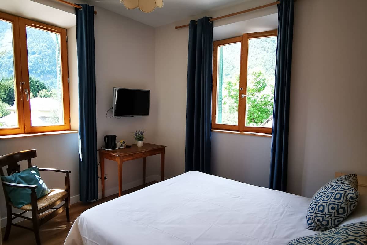 Een slaapkamer met een bed, een bureau, een stoel, een televisie en grote ramen met gordijnen.