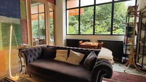 Un salon chaleureux avec un canapé aux couleurs sombres, une grande fenêtre donnant sur un paysage verdoyant et une bibliothèque remplie de livres et d’objets décoratifs.