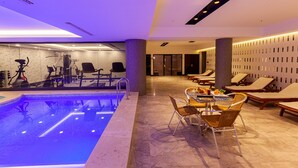 A modern indoor pool area with exercise equipment, lounge chairs, and a dining table. - Ilci Residence Hotel (Ankara)