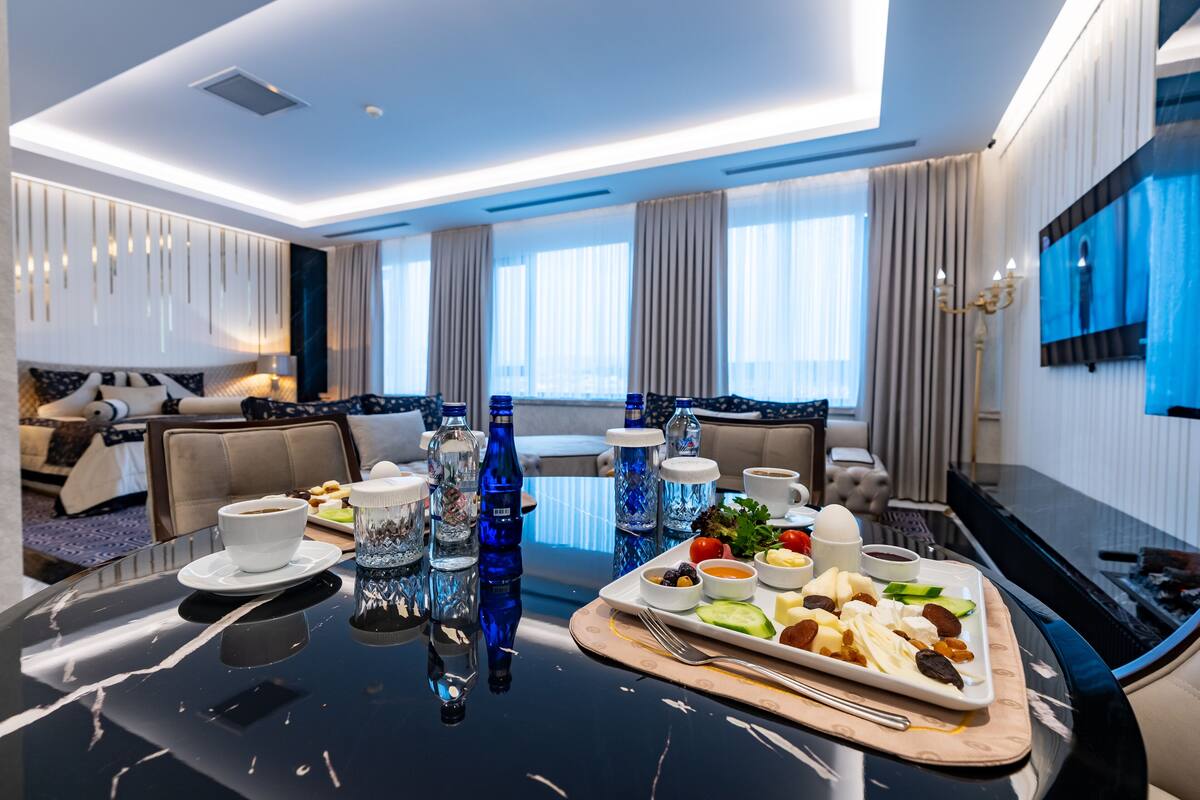 A modern hotel room with a dining table set for breakfast, including plates of food, cups, and bottles.