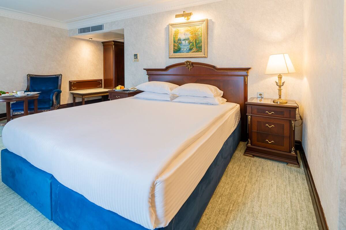 A hotel room with a large bed, a bedside table with a lamp, a chair, and a desk with a telephone.