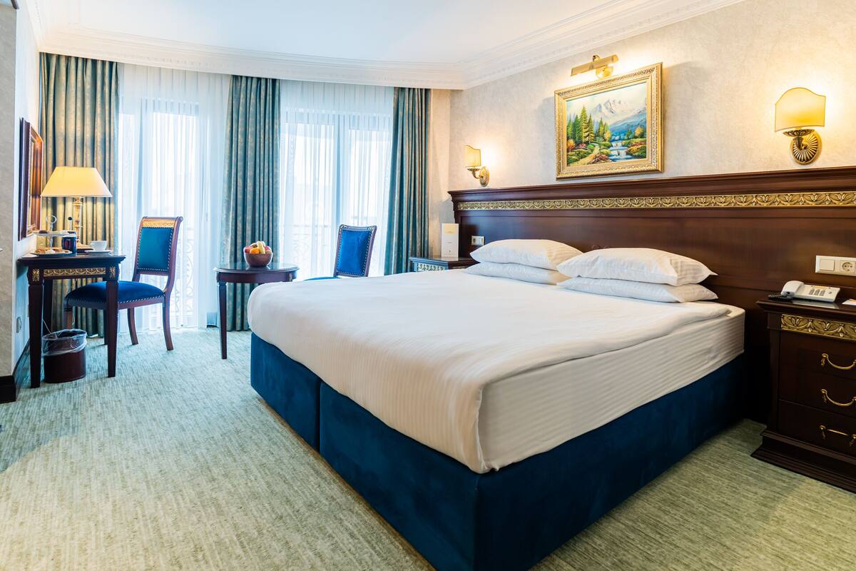 A hotel room with a large bed, a desk with a chair, a small table with a bowl of fruit, and a painting on the wall.