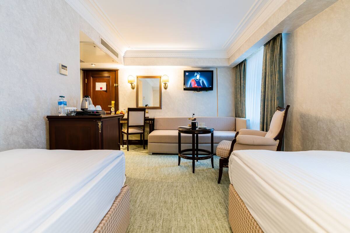 A hotel room with two beds, a desk with a chair, a small table, a TV, and a window with curtains.